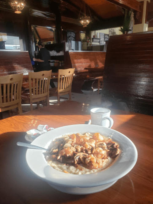 A bowl of oatmeal topped with fruit sits on a wooden table, with a coffee cup and a cozy café background.